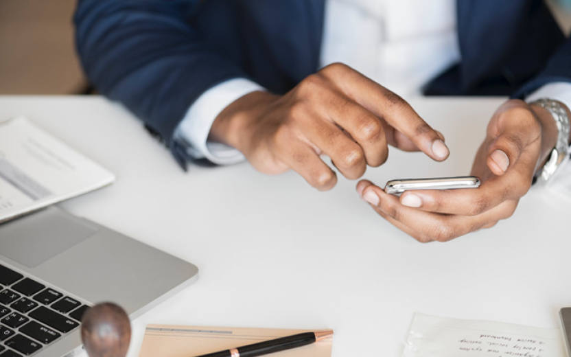 User's hands holding a mobile phone and tapping a code, with a laptop and other items visible on the users desk, and with the Duo logo superimposed on the top left of the image.