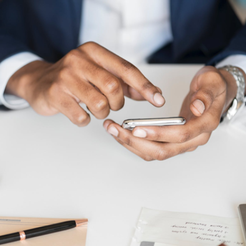User's hands holding a mobile phone and tapping a code, with a laptop and other items visible on the users desk, and with the Duo logo superimposed on the top left of the image.