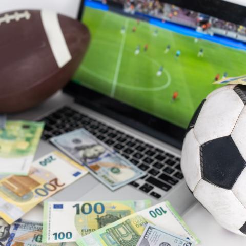 Adobe stock photo of sports balls, cash, and a laptop showing a US football game