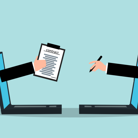 One hand reaches out of a laptop holding a contract, opposite another hand reaching out and holding a pen.